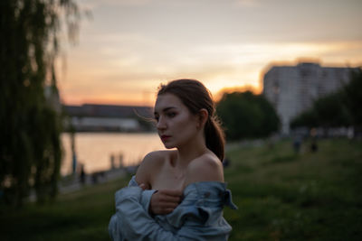 Young woman looking away while standing outdoors during sunset