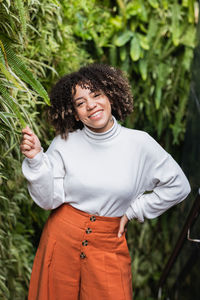Optimistic african american female with curly hair standing near green hedge in park while touching leaf and looking at camera