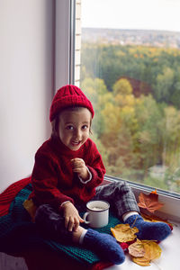 Boy a child in a red sweater and a knitted hat sits at the window on the windowsill