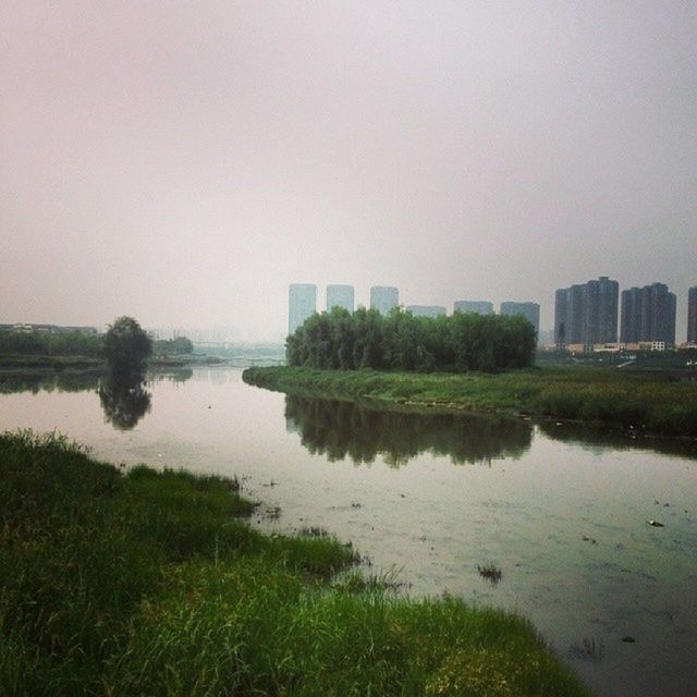 water, grass, clear sky, copy space, lake, reflection, building exterior, built structure, architecture, tranquility, tranquil scene, river, nature, sky, beauty in nature, scenics, plant, riverbank, tree, growth