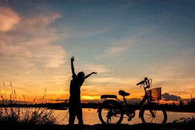 Silhouette people standing by bicycle against sky during sunset