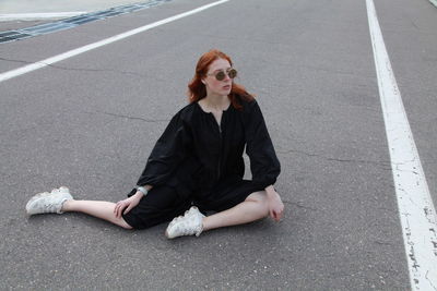 Fashion portrait of ginger woman in loose black dress and sneakers sitting posing on road outside 