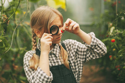 A beautiful little girl stands in a greenhouse with a crop, holds a magnifying glass 