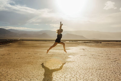 Side view of young woman jumping at desert against sky during sunny day