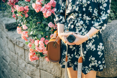 Midsection of woman holding digital camera while standing by flowering plant