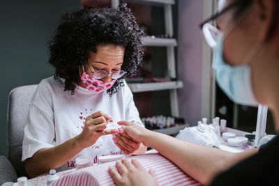 Ethnic female artist applying nail polish on fingernails of client during manicure in beauty studio