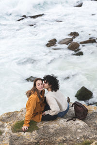Young woman kissing cheek of friend while sitting on cliff by sea