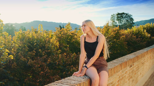 Young woman looking away while sitting on tree against sky