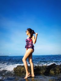Side view of young woman in swimwear standing at beach against blue sky during sunny day