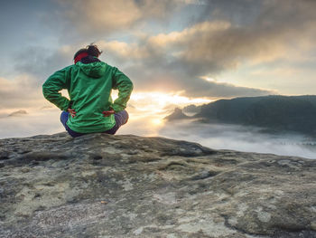 Beautiful girl in a hat and green jacket meditate while sitting on a stone in the mountains