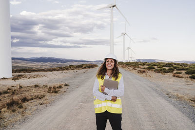 Happy engineer holding laptop standing on dirt road at wind farm