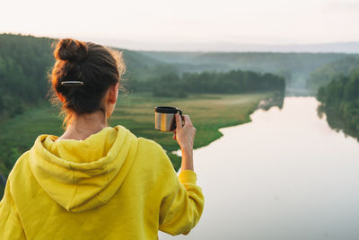 Young woman girl traveler in yellow hoodie with cup of the morning coffee looking at sunrise 