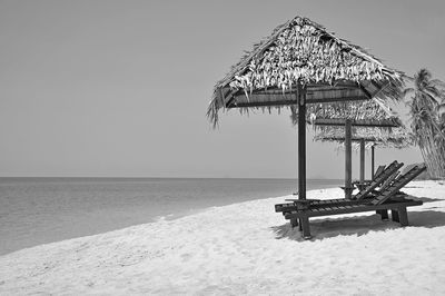 Lounge chairs below thatched roof parasols on shore against clear sky