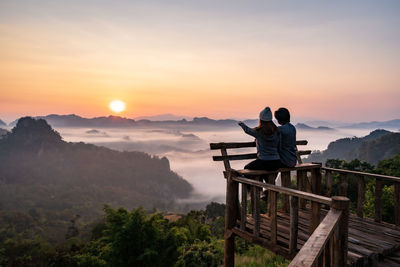 Rear view of people sitting on mountain against sunset sky