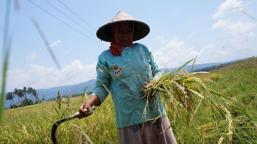 Woman holding sickle while standing in farm against sky