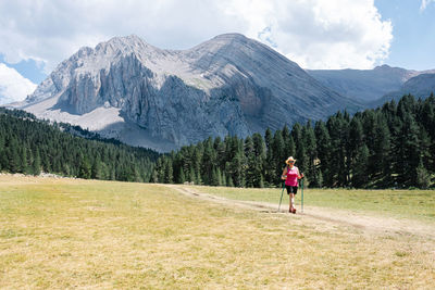 Hiker woman wearing straw hat, shorts and backpack on the path across a plain walking with amazing mountains at the background while enjoys the natural environment around. horizontal photo.