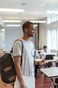 Smiling young student with backpack holding mobile phone while standing in classroom