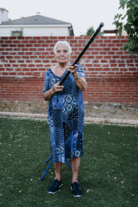 Full length of senior woman holding stick standing against brick wall