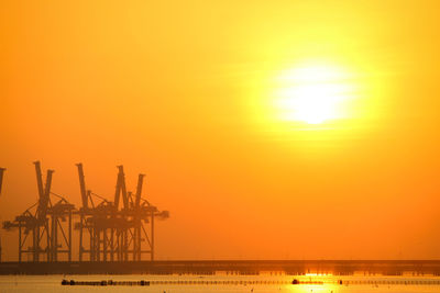 Cranes at harbor against bright sun in sky during sunrise