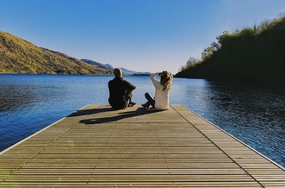 Rear view of couple sitting on jetty over lake against clear sky
