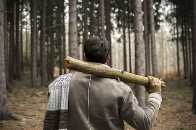 Rear view of young man holding log while standing in forest