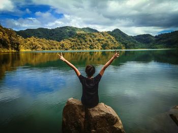 Rear view of woman sitting with arms raised on rock by lake against sky