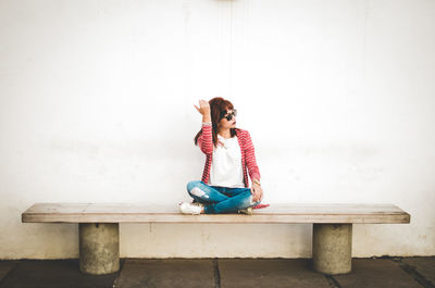 Full length of young woman sitting on bench against wall