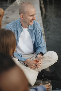 Smiling man holding wineglass while sitting cross-legged on pier