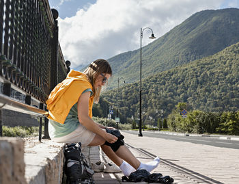 Young woman in yellow putting on protective equipment before roller skating on embankment 