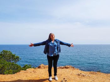 Mature man with arms outstretched standing on cliff by sea against sky