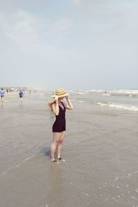 Full length of woman wearing hat standing on shore at beach