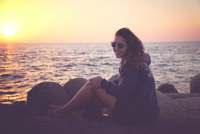 Full length of smiling young woman relaxing on retailing wall by sea during sunset