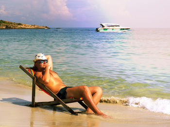 Full length of shirtless man resting on deck chair at beach during summer