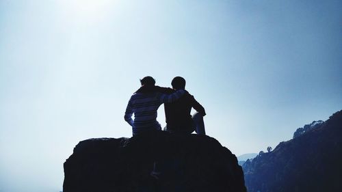 Rear view of men sitting on rock against clear sky during sunny day