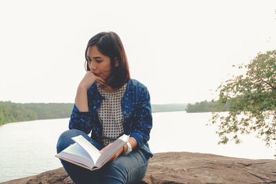 Young woman thinking while holding book on rock by sea against clear sky