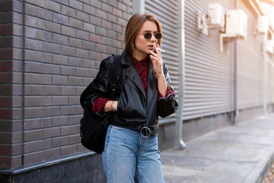 Young woman using phone while standing against wall