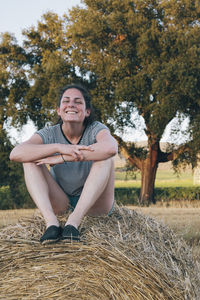 Young woman smiling wide atop a bale of hay on a farm