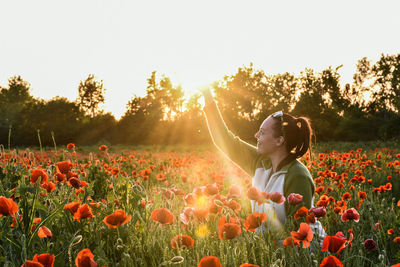 Woman standing by flowering plants on field against sky during sunset