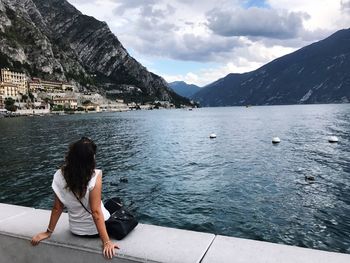 Rear view of woman sitting on retaining wall by lake against sky