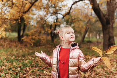 Happy little blonde girl plays with yellow autumn leaves in garden. kid throwing up leaves in park