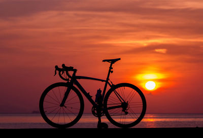 Silhouette bicycle by sea against orange sky