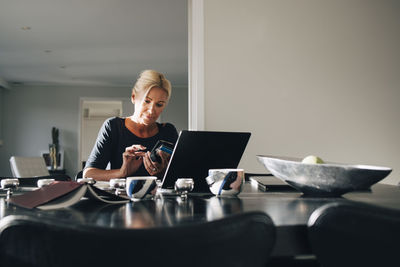Mature blond woman using smart phone with laptop on dining table at home