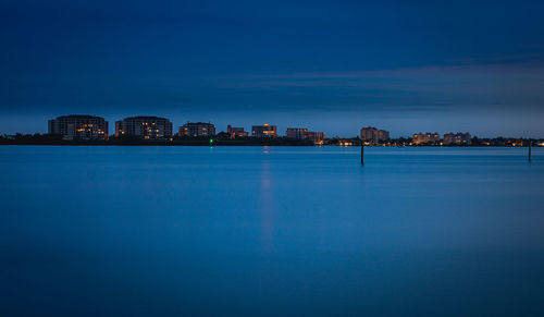 Illuminated buildings by sea against blue sky at dusk