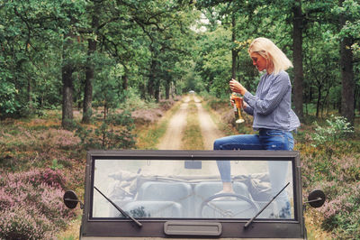 Woman holding bottle while standing in jeep at forest