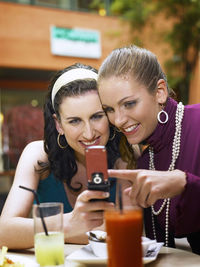 Cheerful female friends using phone while sitting at outdoor cafe