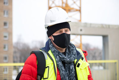 Portrait of thoughtful civil engineer architect wearing hardhat and black face mask at building site