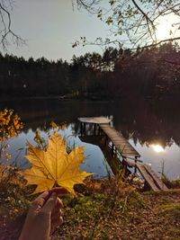 Person holding maple leaves by lake against sky during autumn