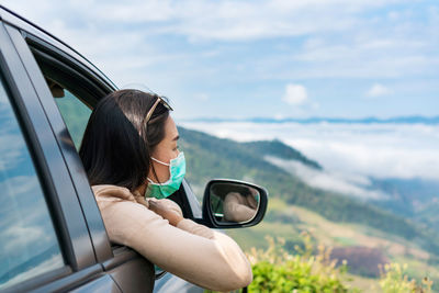 Woman wearing mask leaning out from car window against landscape