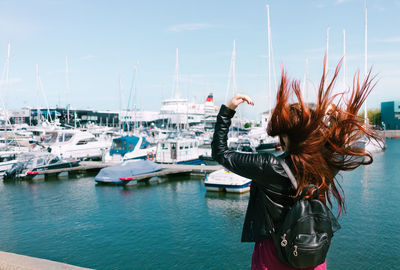 Rear view of young woman tossing hair while standing at harbor against sky