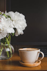 Cozy home workplace. stylish cup coffee, small glass vase with white peonies near computer monitor